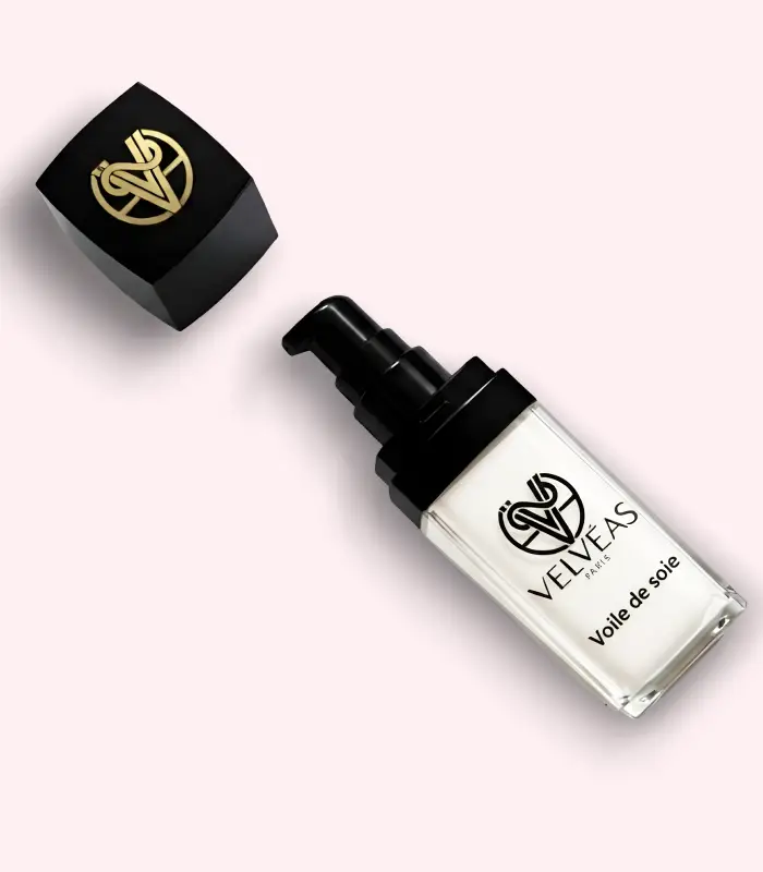 Flacon ouvert Velvéas Voile de Soie avec pompe doseuse noire et capuchon avec logo doré posé à côté sur fond rose pâle