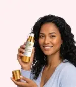 Jeune femme souriante aux cheveux bouclés noirs tenant le flacon ouvert Velvéas Crème Anti-Âge Face 50 ml avec capuchon doré sur fond rose pâle