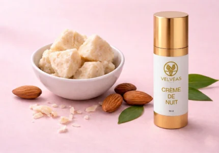 Flacon Velvéas Crème de Nuit 50 ml à côté d'un bol de beurre de karité brut et d'amandes entières sur fond rose pâle