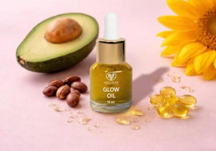 Flacon Velvéas Glow Oil 15 ml entouré d'un demi-avocat, graines de jojoba, capsules de vitamine E et fleur de tournesol sur fond rose pâle